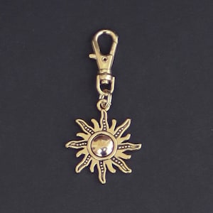 May include: A gold-toned sun keychain with a silver-toned ring and a silver-toned clasp. The sun has a raised center and a textured design.