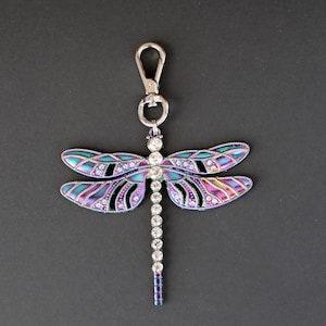 May include: A dragonfly keychain with iridescent wings and a silver clasp. The wings feature a gradient of blue, purple, and pink hues, accented with clear rhinestones. The body is a line of clear rhinestones.