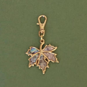 May include: A gold-colored leaf charm with an abalone inlay, attached to a gold-tone lobster clasp. The leaf design features intricate details and a natural, iridescent pattern. The charm is suspended against a green background.