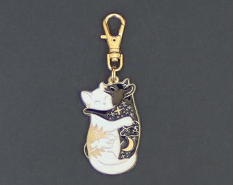 May include: A gold-colored metal keychain featuring two hugging cat figures, one white and one black. The white cat has a sun design, and the black cat has a moon and star design. The keychain has a gold clasp.