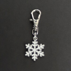 May include: A silver-colored metal keychain with a lobster clasp and a snowflake charm. The snowflake is white with a silver outline. The keychain is displayed against a black background.