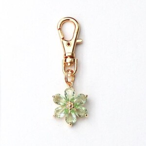 May include: A gold-colored metal clasp with a flower charm. The charm is a six-petal flower with light green gemstone petals and a gold-colored center. The clasp is designed to attach to a keychain or other accessory.