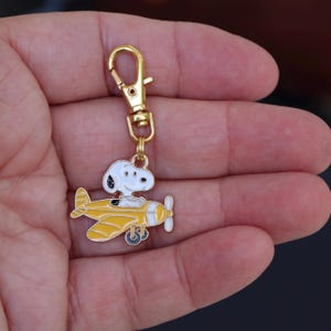 May include: A gold-colored keychain with a cartoon Snoopy character in a yellow airplane. The keychain has a lobster clasp for easy attachment. The Snoopy figure is white with black accents, and the plane has a white propeller.