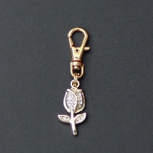 May include: Gold-toned metal keychain with a rose pendant. The rose pendant is encrusted with small, clear crystals. The keychain has a lobster clasp for easy attachment to bags or keys. The background is a solid dark gray.