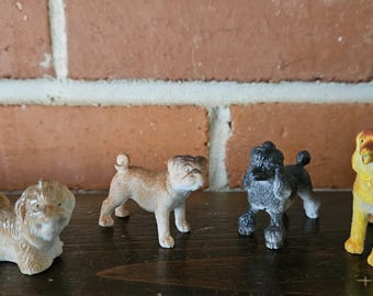 May include: Four small dog figurines in a row. The dogs are a light brown Shih Tzu, a tan pug, a gray poodle, and a yellow and orange lion-like dog. The figurines are on a dark brown surface in front of a brick wall.