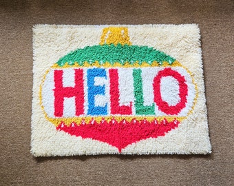 May include: A white, fluffy rug with a colorful, embroidered ornament in the shape of a Christmas tree. The ornament says "HELLO" in red, blue, green, and yellow letters.