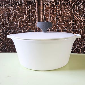 May include: A white enameled cast iron Dutch oven with a lid and a dark gray handle. The pot has two side handles and sits on a light green surface. The background is a woven brown pattern.
