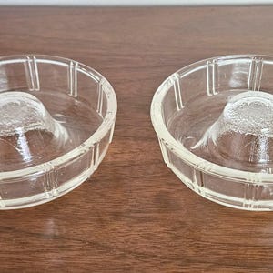 May include: Two clear glass citrus juicers, each with a central reamer and a ridged outer bowl. The juicers are displayed on a dark wooden surface, showcasing their transparent design and functional form.