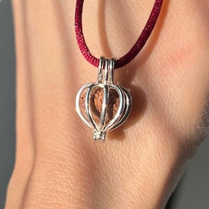 Peut inclure: Un pendentif en argent en forme de cœur avec une graine brune à l'intérieur, suspendu à un cordon rouge.