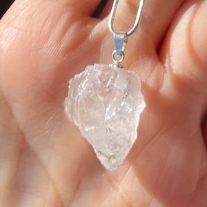 Può includere: Un ciondolo in cristallo di quarzo trasparente su una catena d'argento. Il cristallo è grezzo e ha una forma naturale.