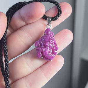 Könnte beinhalten: Ein rosa Edelstein-Anhänger mit einem detaillierten Design einer Gottheit, der an einer schwarzen Schnurkette hängt.