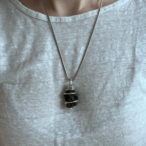 May include: A silver necklace with a dark, irregularly shaped pendant. The pendant is wrapped in silver wire. The necklace is worn against a light-colored shirt.