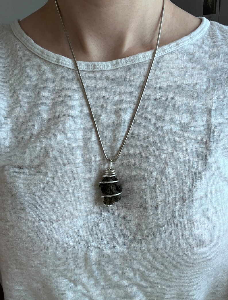 May include: A silver necklace with a dark, irregularly shaped pendant. The pendant is wrapped in silver wire. The necklace is worn against a light-colored shirt.