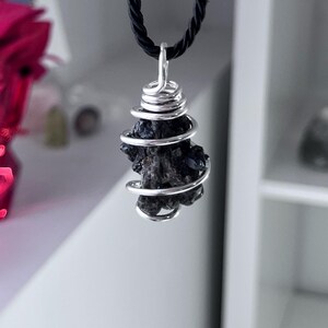 May include: A dark, textured stone pendant wrapped in silver-colored wire, suspended from a black cord. The stone has a rough, irregular shape. The wire is coiled around the stone. The necklace is displayed against a neutral background.