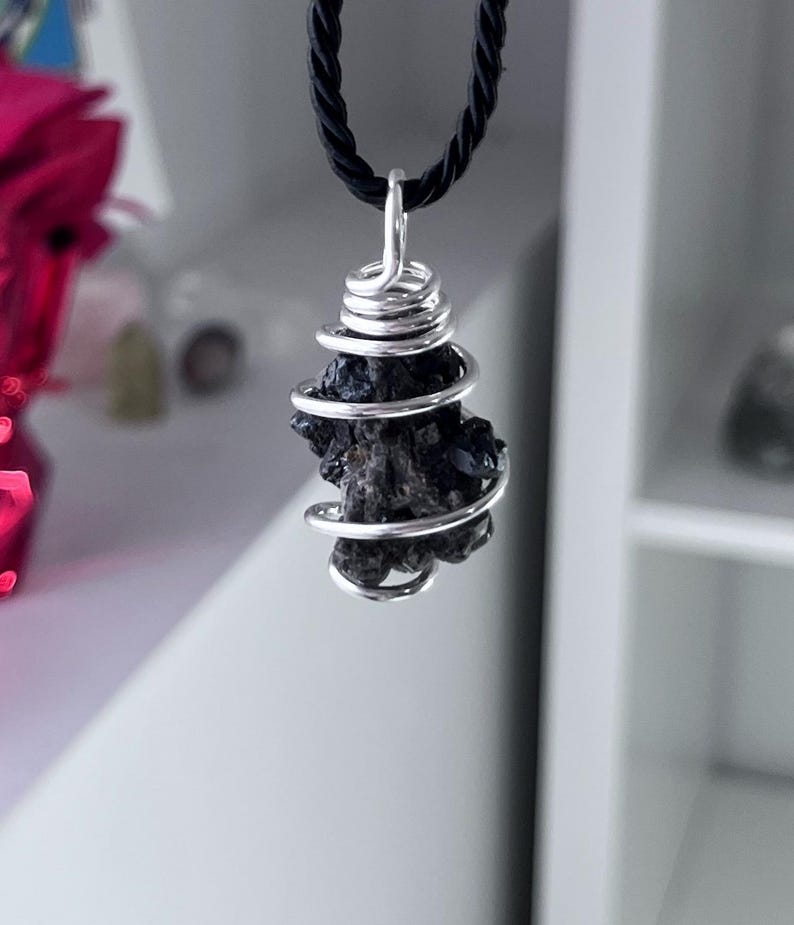 May include: A dark, textured stone pendant wrapped in silver-colored wire, suspended from a black cord. The stone has a rough, irregular shape. The wire is coiled around the stone. The necklace is displayed against a neutral background.