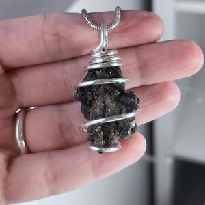 May include: A dark, rough-textured stone pendant wrapped in silver-colored wire, suspended from a silver chain. The stone has a natural, irregular shape and a dark, almost black color. The wire is tightly coiled around the stone.