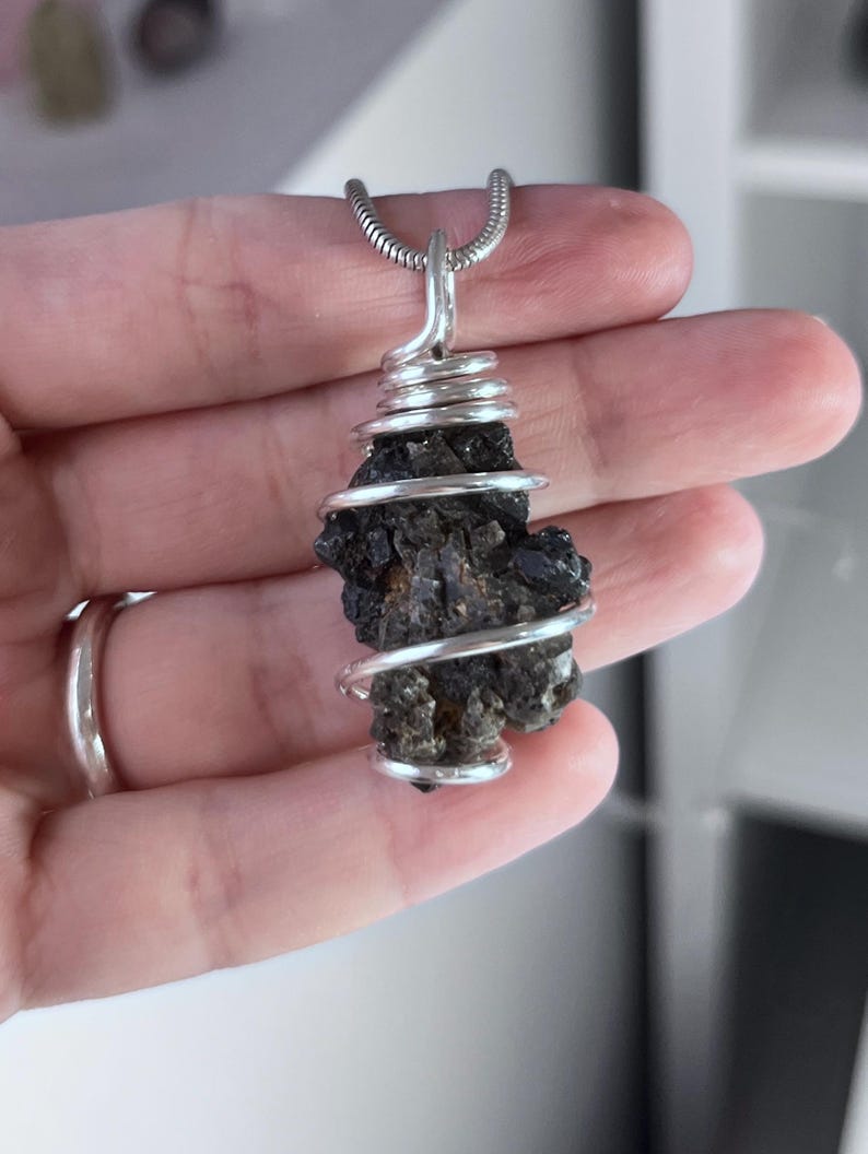 May include: A dark, rough-textured stone pendant wrapped in silver-colored wire, suspended from a silver chain. The stone has a natural, irregular shape and a dark, almost black color. The wire is tightly coiled around the stone.