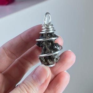May include: A dark, irregularly shaped pendant wrapped in silver wire. The wire is coiled around the stone and forms a loop at the top for hanging. The pendant is held in a hand.
