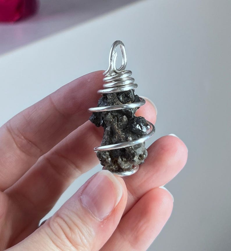 May include: A dark, irregularly shaped pendant wrapped in silver wire. The wire is coiled around the stone and forms a loop at the top for hanging. The pendant is held in a hand.