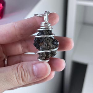 May include: A silver wire-wrapped pendant featuring a dark, textured stone. The stone is a deep brown color with a rough, natural appearance. The wire is coiled around the stone and forms a loop for a chain.