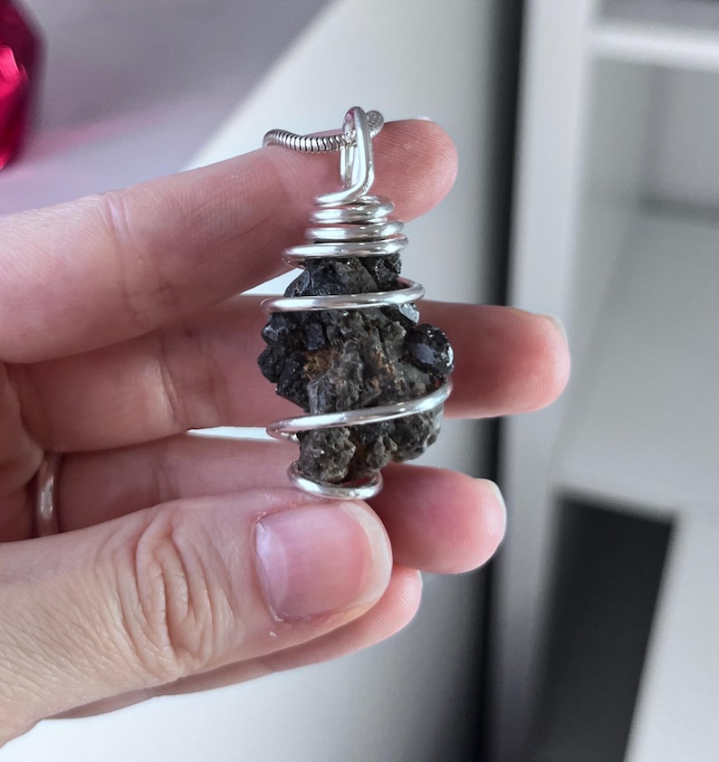 May include: A silver wire-wrapped pendant featuring a dark, textured stone. The stone is a deep brown color with a rough, natural appearance. The wire is coiled around the stone and forms a loop for a chain.