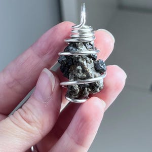 May include: A dark, irregularly shaped stone pendant wrapped in silver-colored wire. The wire is coiled around the stone and forms a loop at the top for hanging. The stone has a rough, textured surface.