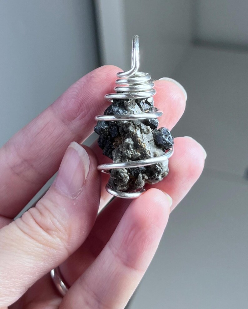 May include: A dark, irregularly shaped stone pendant wrapped in silver-colored wire. The wire is coiled around the stone and forms a loop at the top for hanging. The stone has a rough, textured surface.