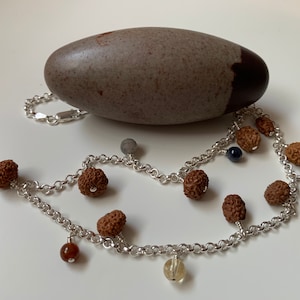 Può includere: Una collana a catena d'argento con perline marroni, perline d'argento e perline di pietre preziose colorate. La collana è appoggiata su una superficie bianca accanto a una grande pietra ovale di colore grigio e marrone.