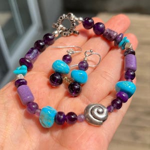 May include: A beaded bracelet and matching earrings set. The bracelet and earrings feature a combination of purple, turquoise, and silver-colored beads. The bracelet has a silver-colored clasp and a decorative silver spiral charm.