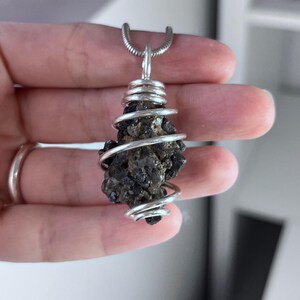 May include: A dark, irregularly shaped pendant with a silver wire wrap. The pendant is suspended from a silver chain. The stone appears to be a dark, possibly volcanic rock, with a rough texture. The wire is tightly wound around the stone.