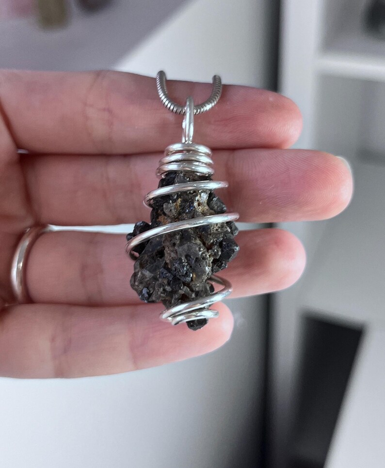 May include: A dark, irregularly shaped pendant with a silver wire wrap. The pendant is suspended from a silver chain. The stone appears to be a dark, possibly volcanic rock, with a rough texture. The wire is tightly wound around the stone.