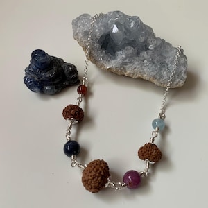 Könnte beinhalten: Eine silberne Halskette mit verschiedenen Perlen, darunter braune, rote, blaue und violette Edelsteine. Die Halskette wird mit einem blauen Kristall und einer dunkelblauen geschnitzten Figur präsentiert.