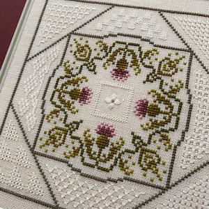May include: A white fabric with a cross-stitch design featuring a green and pink floral pattern. The design is surrounded by a white border with a geometric pattern.