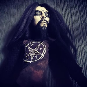 May include: A black and white doll with long black hair and a pentagram on its shirt. The doll is wearing a dark brown shirt with a white pentagram and a goat's head inside. The doll has white eyes and a black beard.