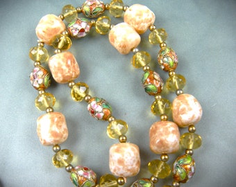 Puede incluir: Un collar largo con cuentas alternas de color marrón claro, amarillo y esmalte multicolor. Las cuentas de esmalte tienen diseños florales en tonos de verde, rosa y blanco.