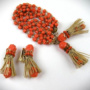 May include: A set of coral and gold tone jewelry featuring a necklace and earrings. The necklace is a long strand of coral beads with gold tone accents and two tassel pendants. The earrings are clip-on style with coral beads and gold tone tassels.