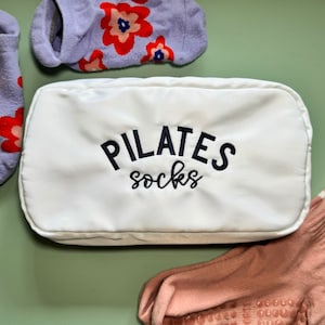 May include: White canvas bag with the text "PILATES socks" embroidered in navy blue. The bag is filled with two pairs of socks, one pair is purple with red flowers and the other pair is pink with a textured grip.