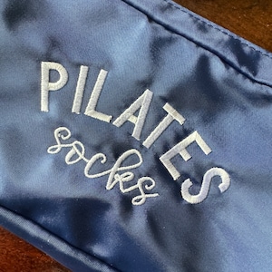 May include: A navy blue zippered pouch with white embroidery that reads "PILATES socks".