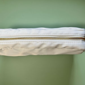 May include: A white zippered pouch with a gold zipper. The pouch is made of a soft, fabric material.
