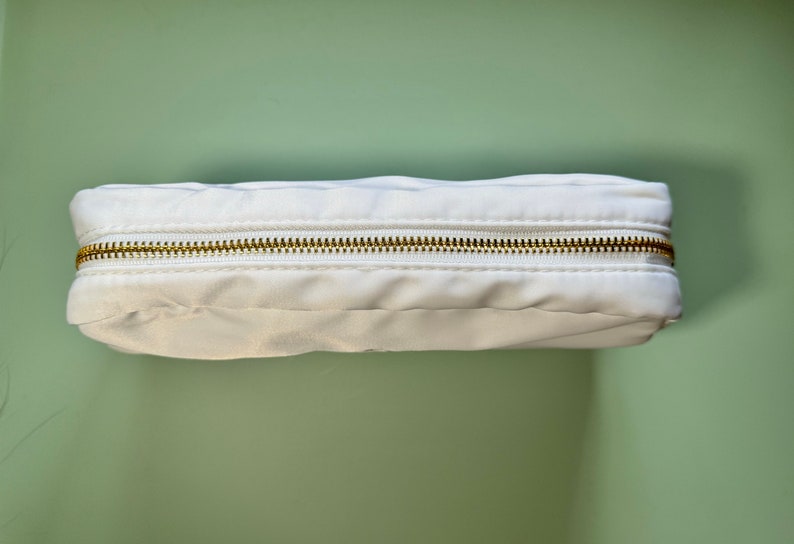 May include: A white zippered pouch with a gold zipper. The pouch is made of a soft, fabric material.
