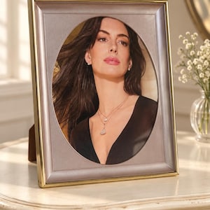 May include: A rectangular picture frame with a metallic gold border and a smooth, mauve-colored inner frame featuring an oval-shaped cutout. The frame displays a photograph of a person with long dark hair wearing a black top and a layered necklace.