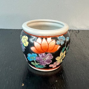 May include: A small, round ceramic planter with a black background and colorful floral design. The planter features a large orange lotus flower surrounded by smaller pink, purple, yellow, and blue flowers.