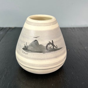 May include: A small, white ceramic vase with gray stripes and a hand-painted scene of a hill, a wheel, and a bird. The vase is signed "Rose".