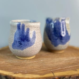 May include: Two small ceramic cups with blue and white abstract designs, sitting on a wooden slice.