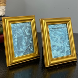 May include: Two gold picture frames with a blue patterned design inside. The frames are sitting on a black surface.