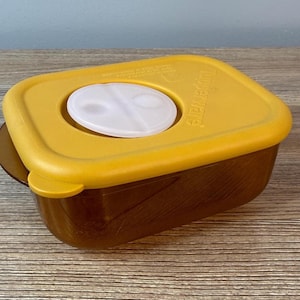 May include: A vintage Tupperware container with a mustard yellow lid and a brown base. The lid features a white, oval-shaped vent. The container is rectangular with rounded corners and is sitting on a wooden surface.