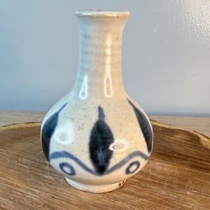 May include: A small, beige ceramic vase with a narrow neck and rounded body. The vase is adorned with dark blue, stylized leaf and swirl patterns. The vase sits on a wooden slice, showcasing its handcrafted design.
