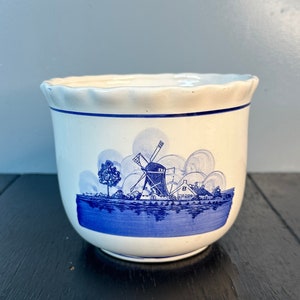 May include: A white ceramic planter with a blue and white windmill scene. The planter has a scalloped rim and a blue and white design of a windmill, houses, and trees.