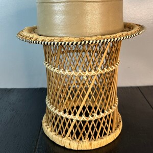May include: A round, light brown planter sits on top of a woven wicker stand with black trim. The stand has a cylindrical shape and is made of natural materials.