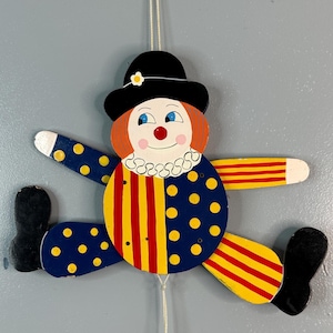 May include: A wooden clown puppet with red and yellow striped pants, blue and yellow polka dot shirt, and a black hat with a white flower. The puppet is hanging from a string and has a red nose and blue eyes.